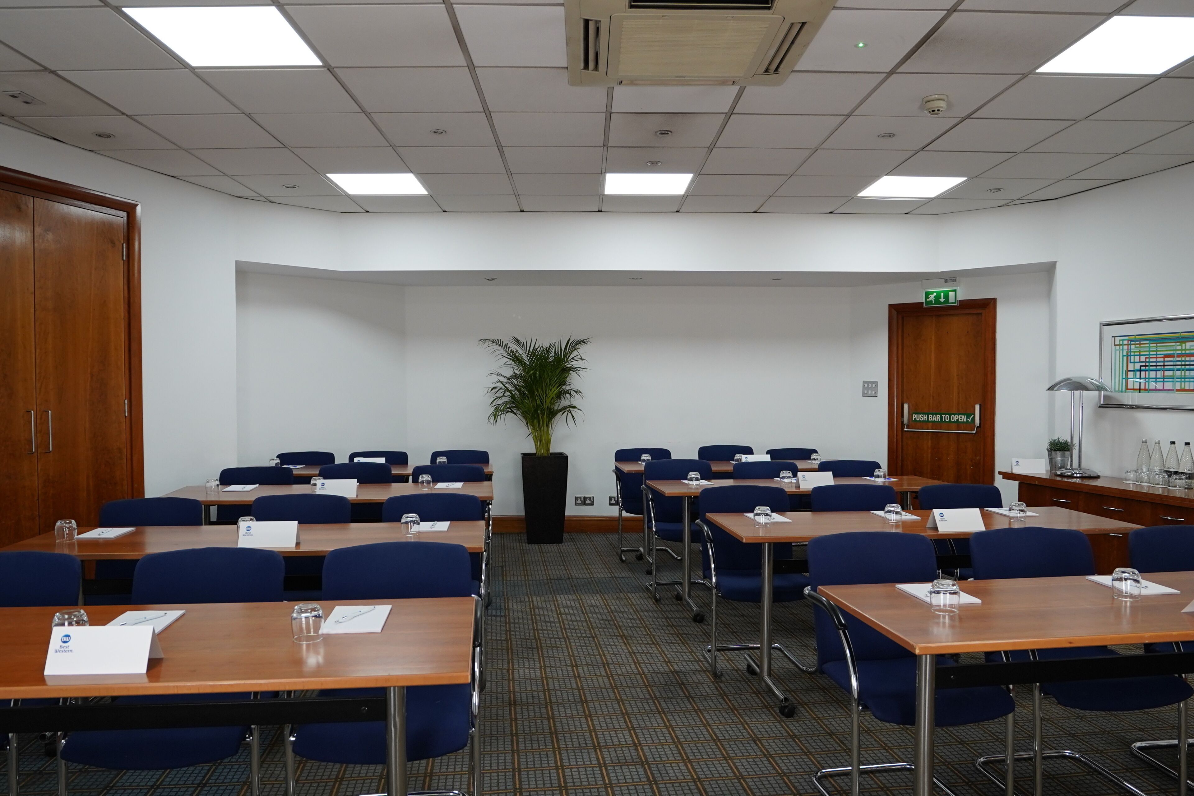 A conference room with tables, chairs, a potted plant, and a push bar to open a door.