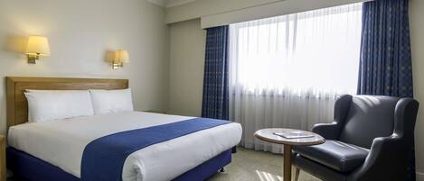 A hotel room with a large bed, a small round table, a leather armchair, and two wall-mounted lamps.