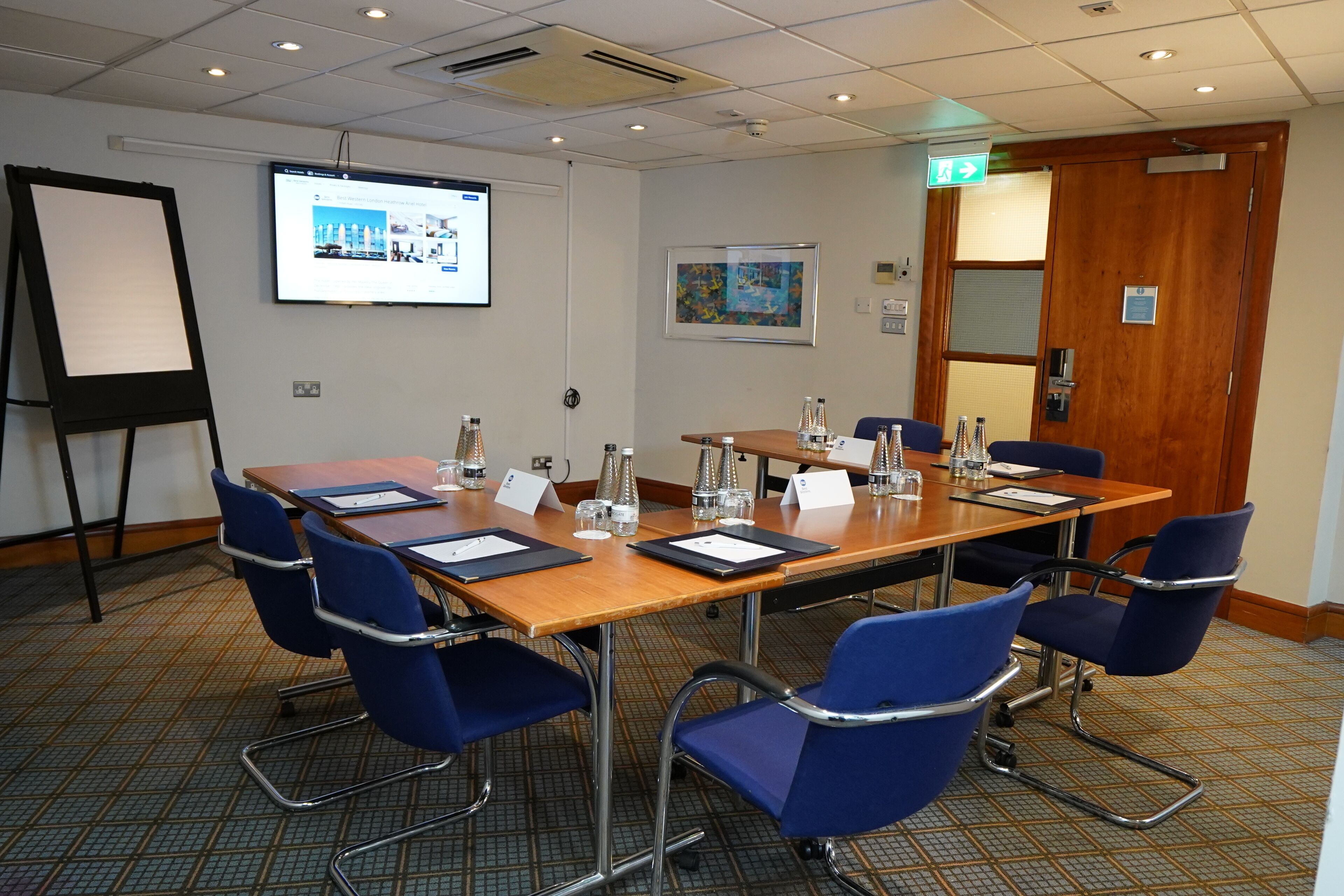 Ett konferensrum med ett långt bord, blå stolar, en projektorduk och en dörr med en utrymningsskylt.