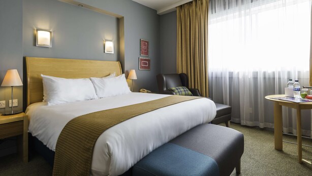 Hotellihuone, jossa on suuri sänky, yöpöydällä lamput, työpöytä tuolilla, pieni pöytä, jonka päällä on pullo ja lasi, sekä ikkuna, jossa on verhot.
