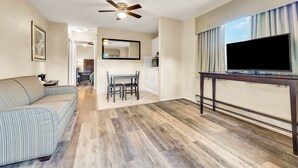 A spacious living room with a large flat-screen TV, a ceiling fan, a dining area with a table and chairs, and a kitchen in the background. - Scottish Fairway Inn (Monterey)