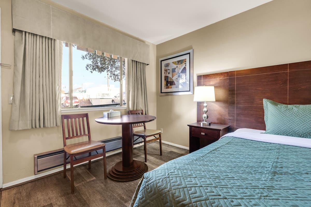 A hotel room with a bed, a desk with a chair, a window with curtains, a wall-mounted lamp, and a framed picture.