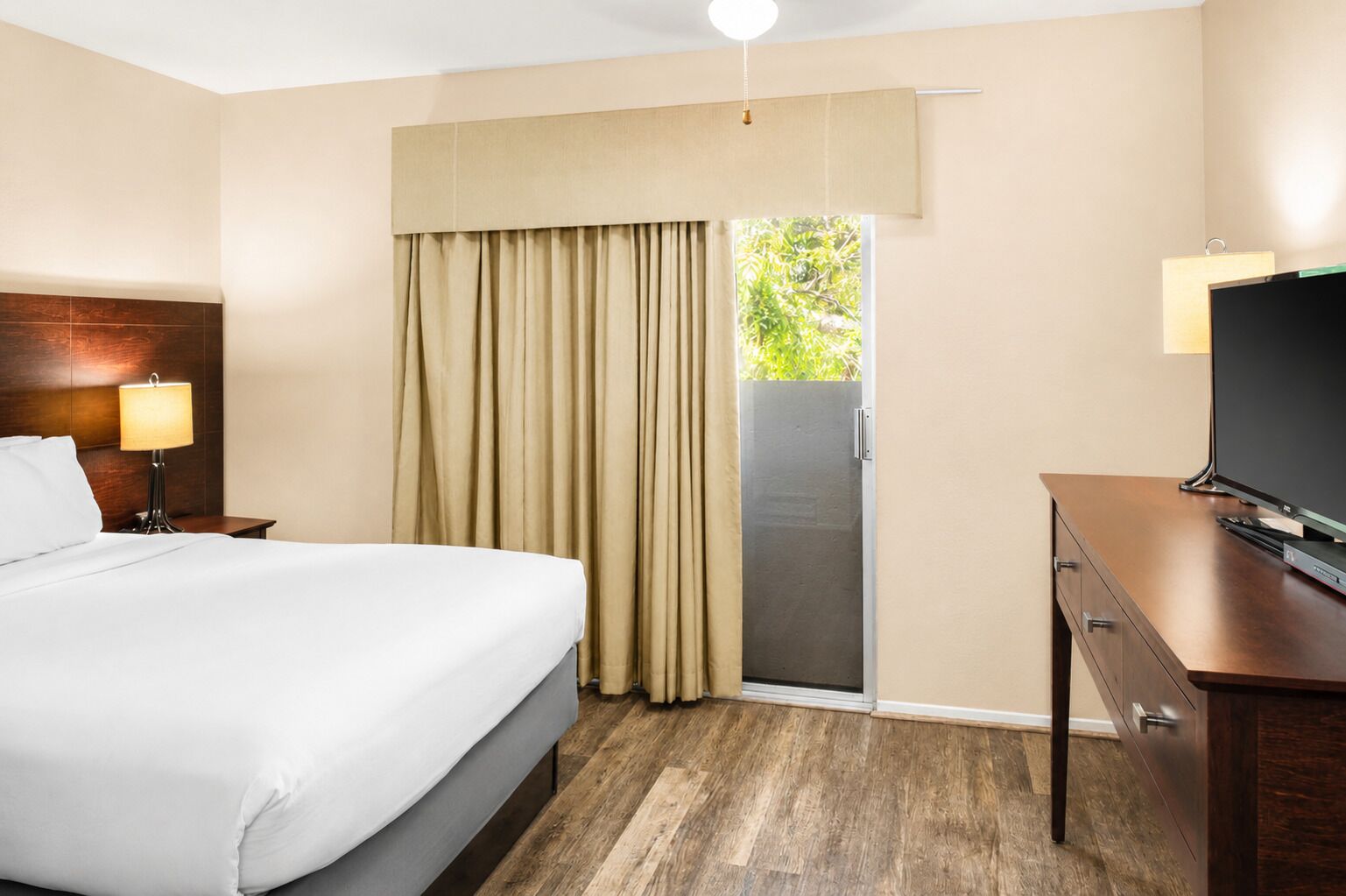 Een hotelkamer met een bed, een bureau met een televisie, een lamp en een schuifdeur naar buiten.