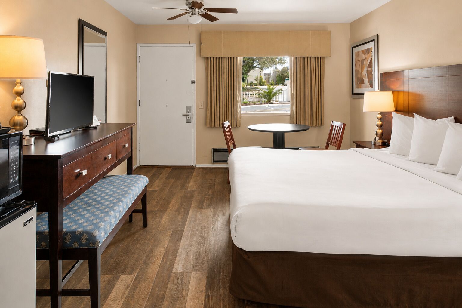 A hotel room with a bed, a desk with a TV, a chair, a small table, and a window with curtains.