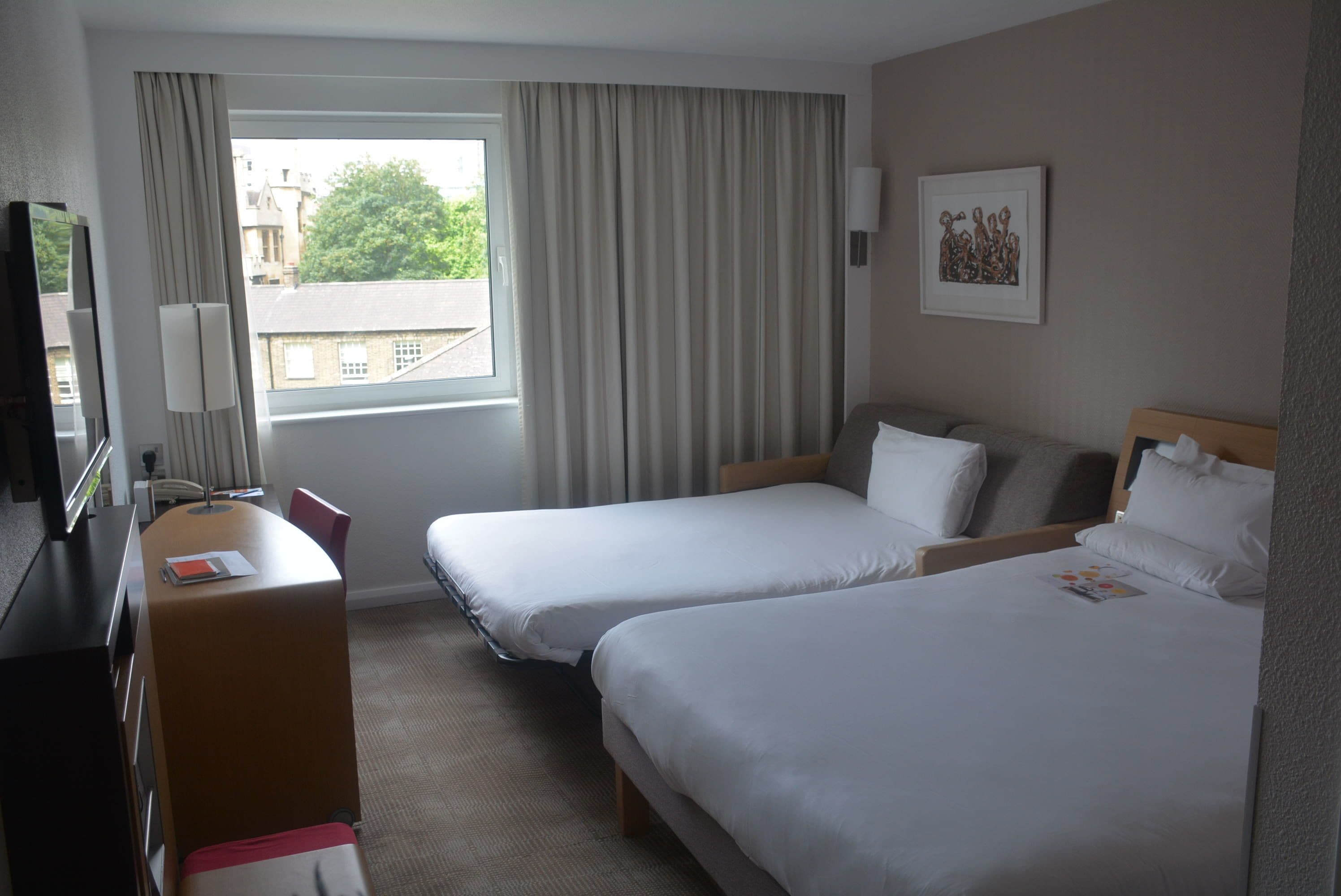 Une chambre d’hôtel avec deux lits simples, un bureau avec une chaise, une télévision et une fenêtre avec des rideaux.