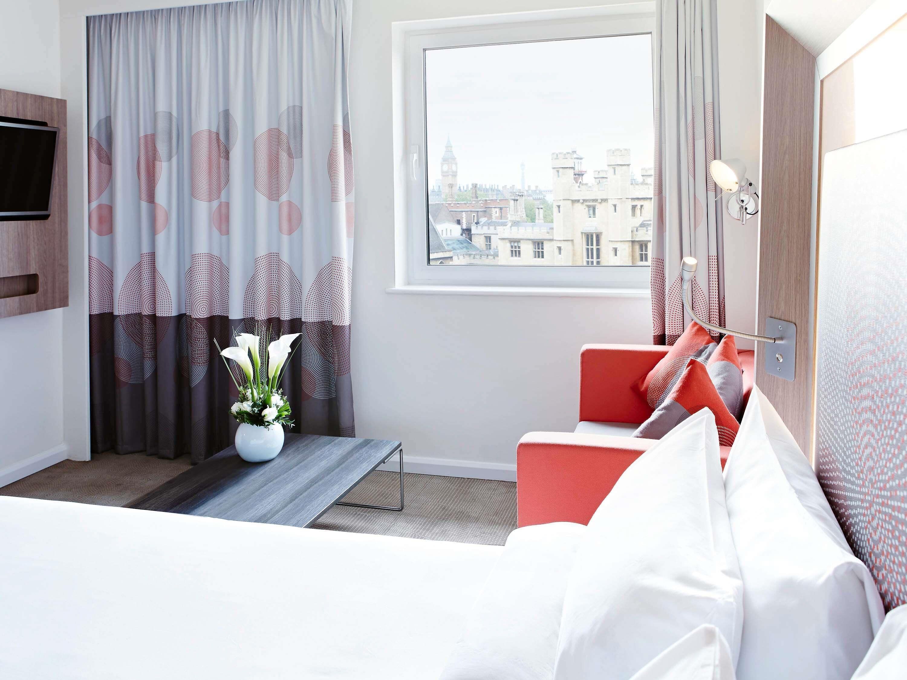 Quarto de hotel com uma cama, uma cadeira vermelha, uma mesinha com um vaso de flores, uma janela com vista para a cidade e uma TV fixada na parede.
