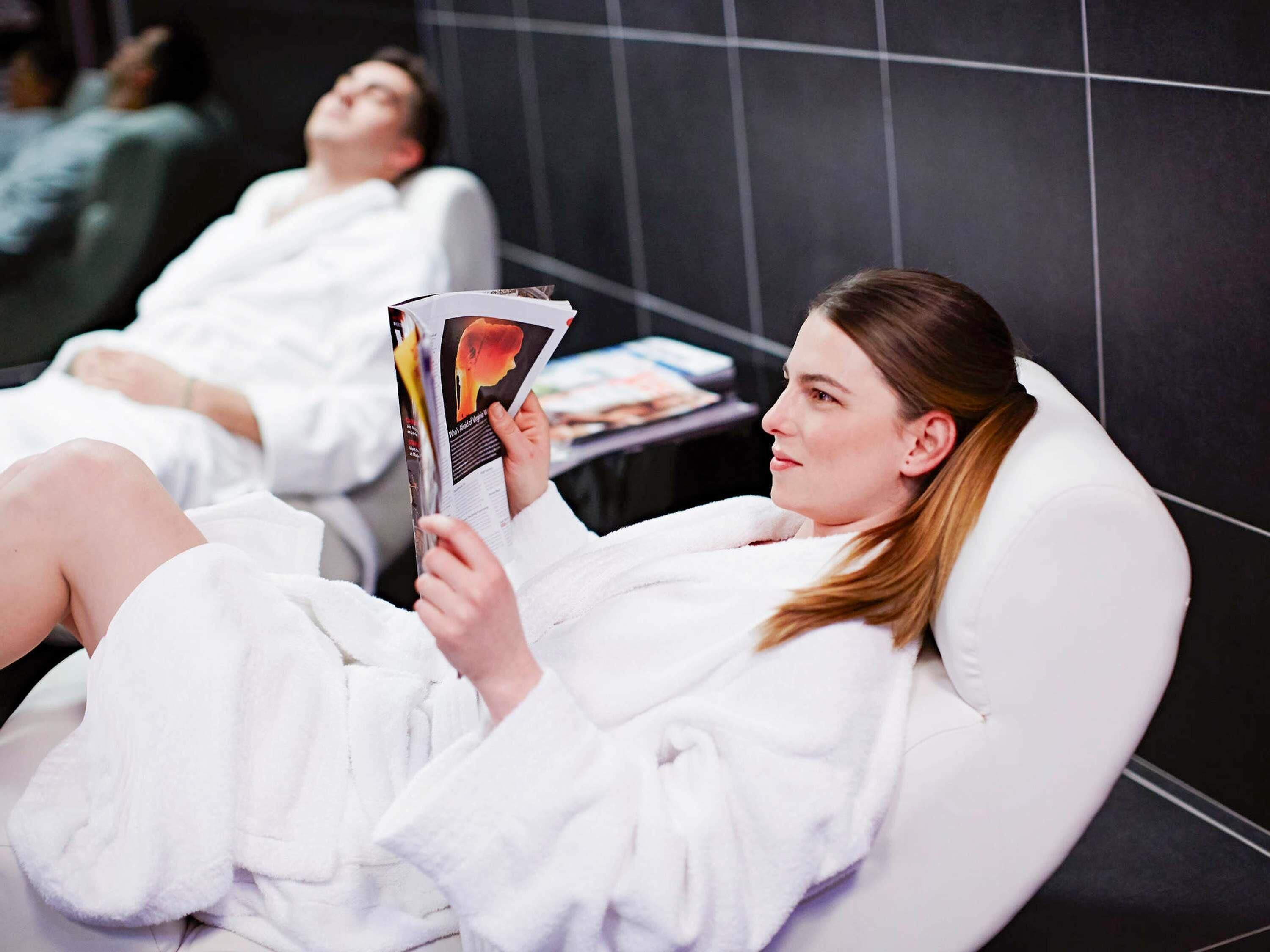 Eine Person liegt entspannt in einem weißen Massagestuhl in einer Wellnessumgebung und liest eine Zeitschrift.