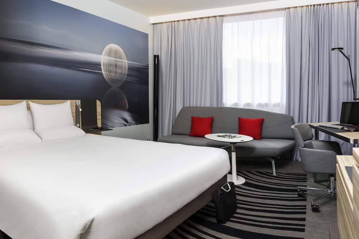 A hotel room with a bed, a sofa, a desk, and a large window with curtains.