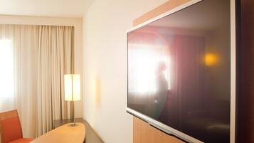Et hotelværelse med et vægmonteret fladskærms-tv, et træbord, en rød stol og en lampe.