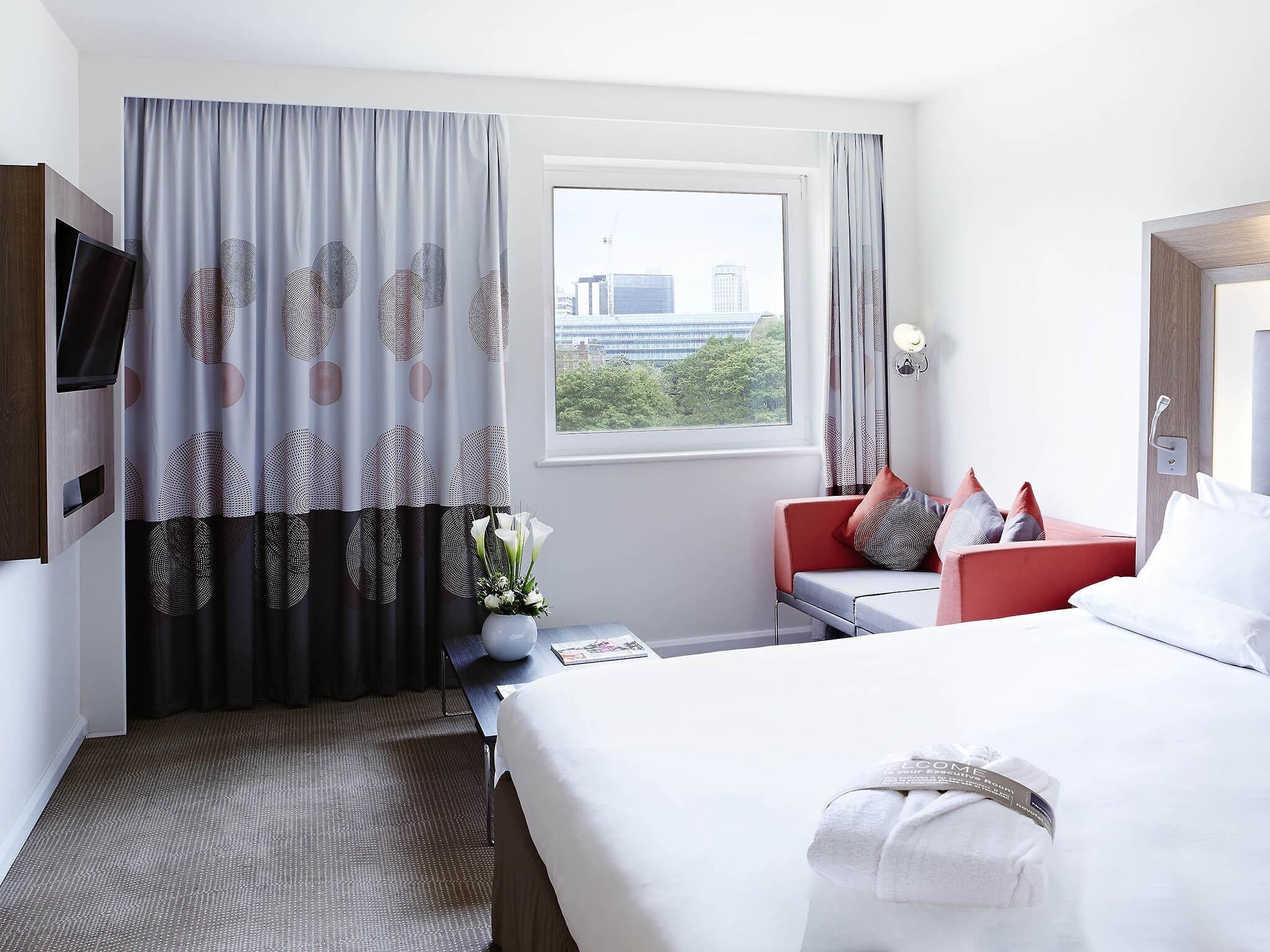 A hotel room with a bed, a red chair, a small table, a TV, and a window with a city view.