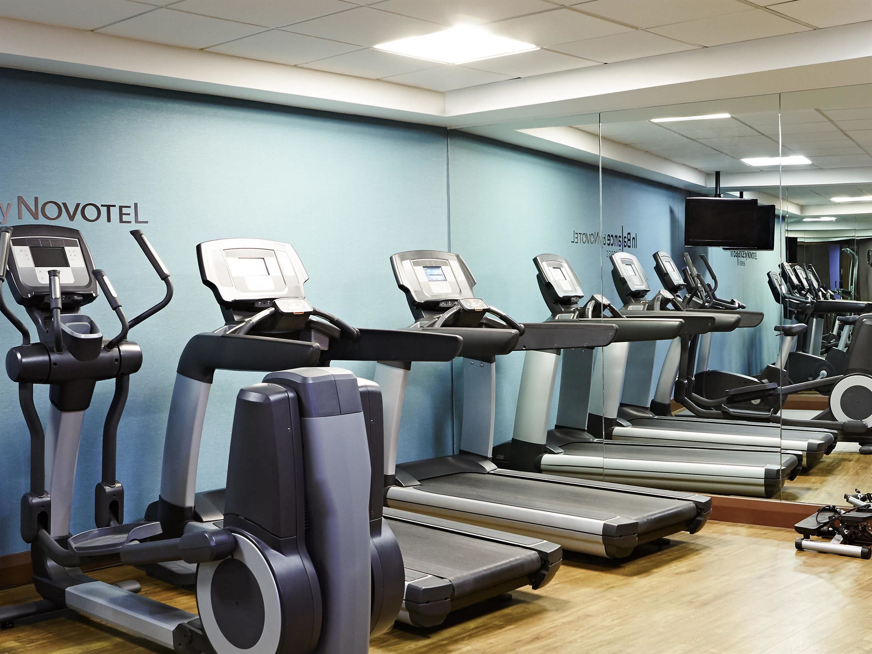 A gym with multiple treadmills and elliptical machines, a wooden floor, and a blue wall with the Novotel logo.
