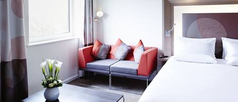 A modern hotel room with a grey sofa, white bedding, and a vase of white flowers.