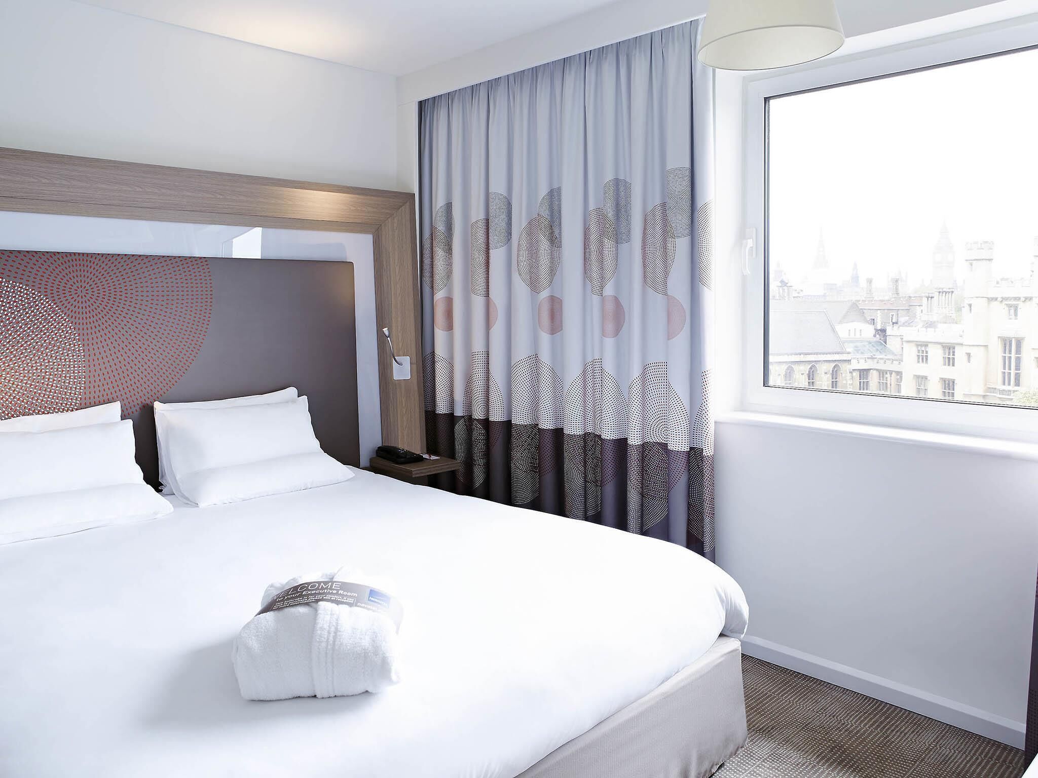 A hotel room with a large bed, a headboard with a geometric pattern, a nightstand, a lamp, and a view of a cityscape through a window.