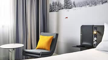 Hotelkamer met een bed, een stoel met geel kussen, een kleine tafel, een aan de muur bevestigde televisie, en een muur met een besneeerd landschap en een vos sticker.