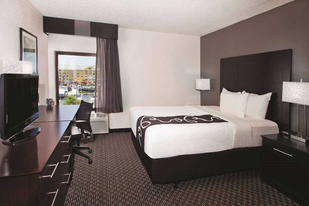 A hotel room with a large bed, a desk with a TV, a chair, and a view of a parking lot and buildings outside.