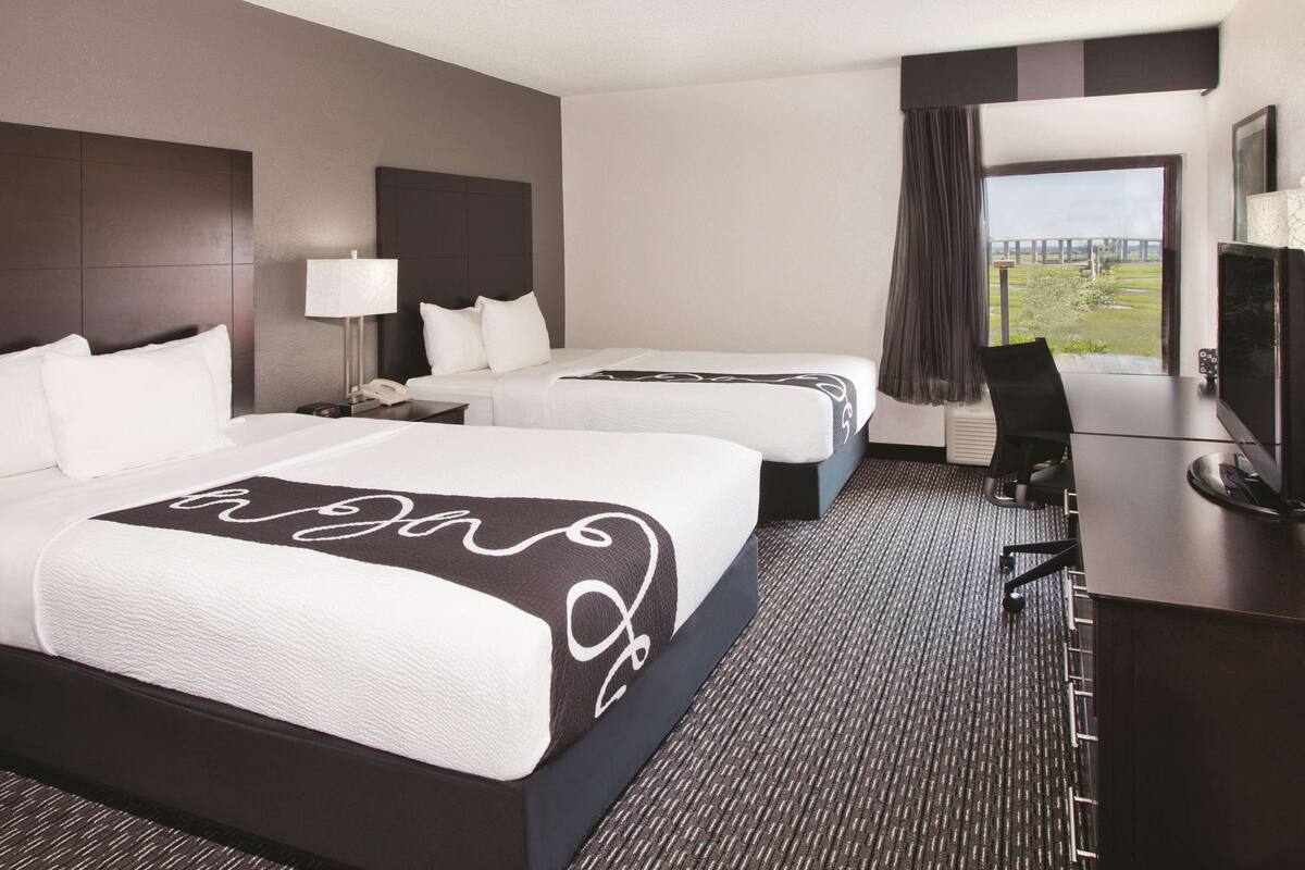 A hotel room with two beds, a desk with a chair, a TV, and a window with a view of a bridge.