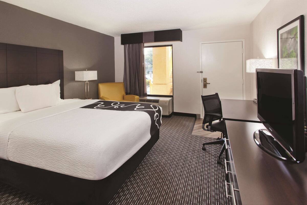 A hotel room with a large bed, a desk with a flat-screen TV, a chair, a lamp, and a window with curtains.