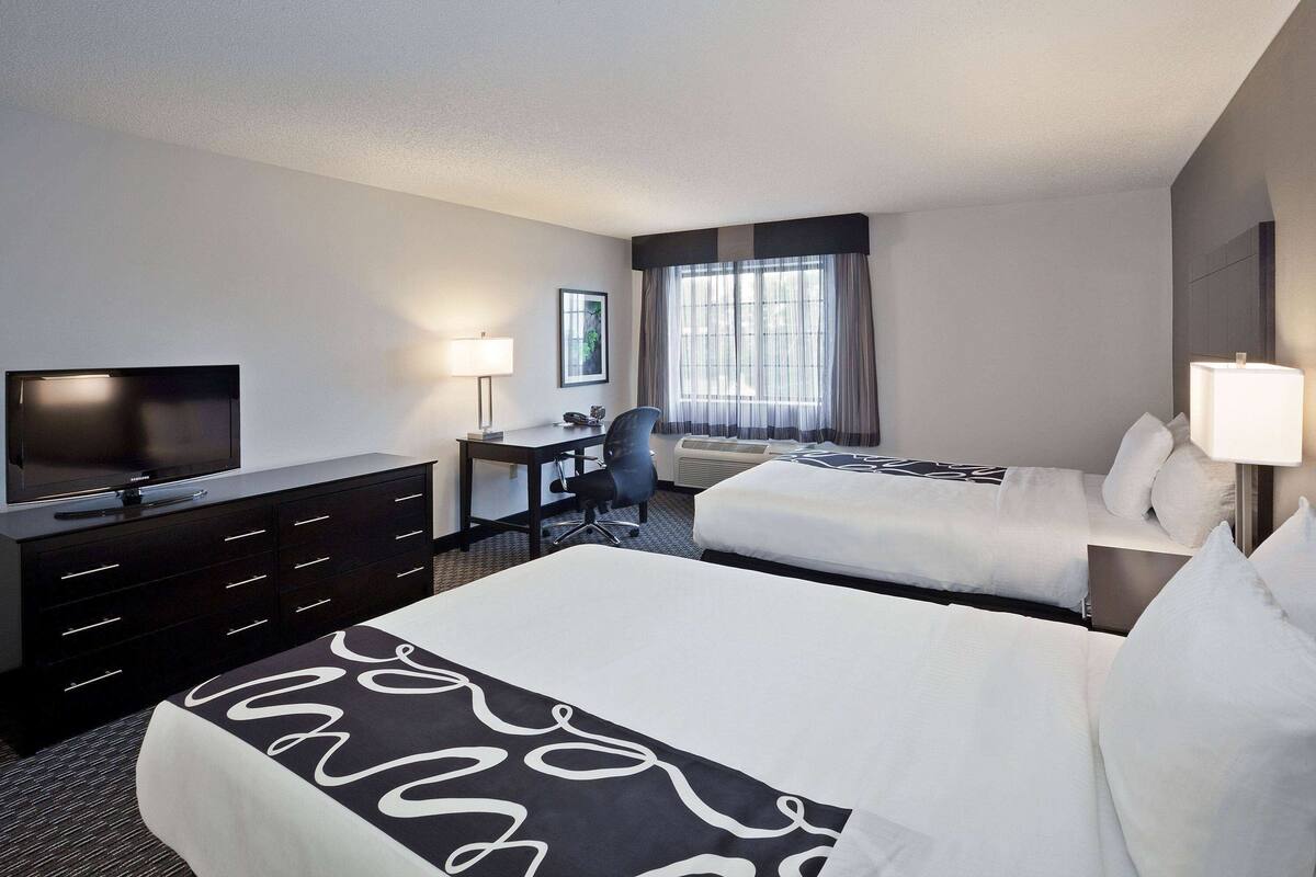 A hotel room with two beds, a desk, a chair, a television, and a large window with curtains.