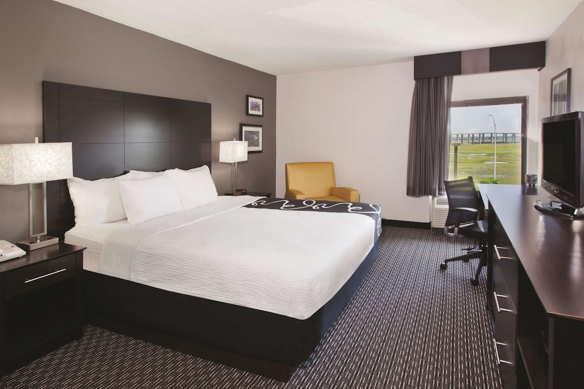 A modern hotel room with a large bed, a desk, a chair, a TV, and a view of a bridge.