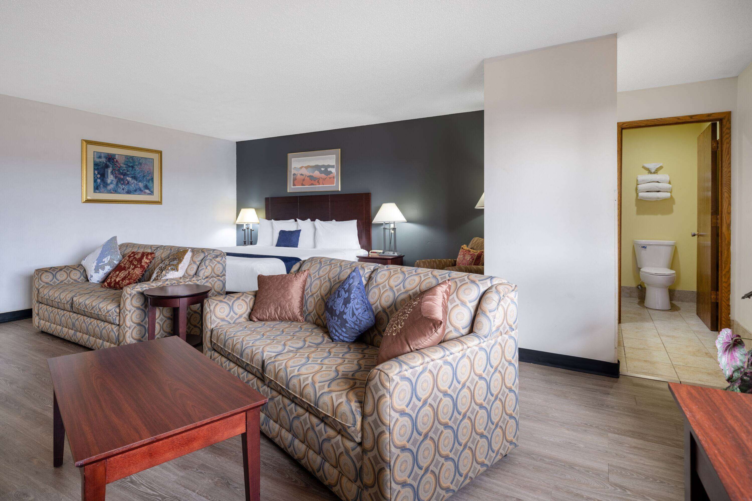Habitación de hotel con un sofá estampado, una mesa de centro de madera, una cama con cabecera, dos lámparas de noche y un baño visible a través de una puerta abierta.