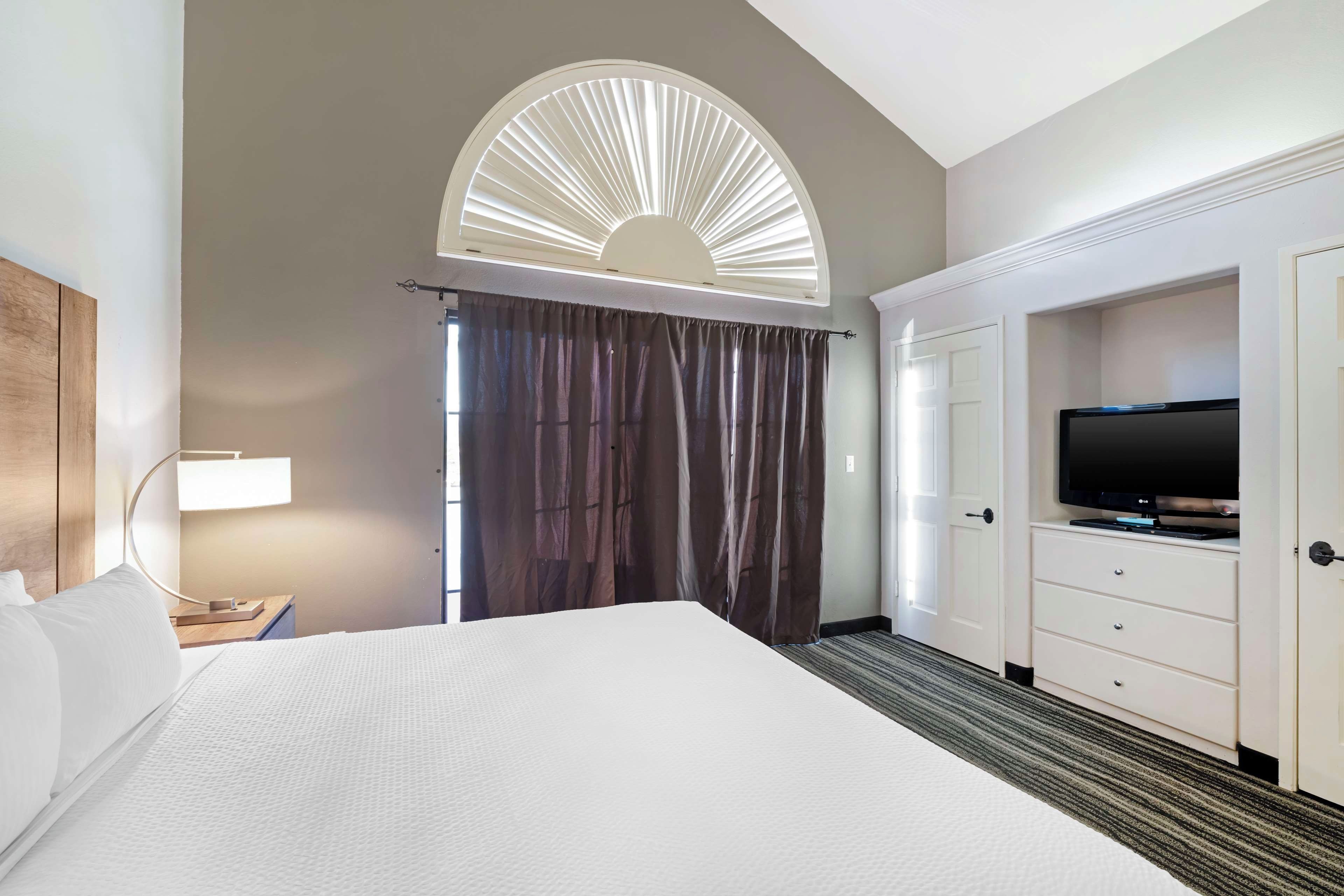 A hotel room with a bed, a TV, a desk with a lamp, and a large window with curtains.
