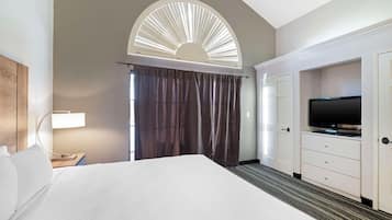A hotel room with a bed, a TV, a desk with a lamp, and a large window with curtains.