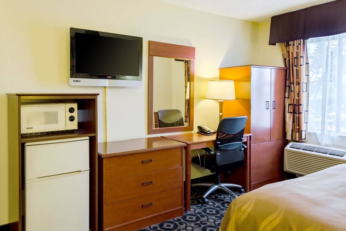 Habitación de hotel con cama, escritorio y silla, microondas, refrigerador, televisor, espejo, lámpara, armario y equipo de aire acondicionado.