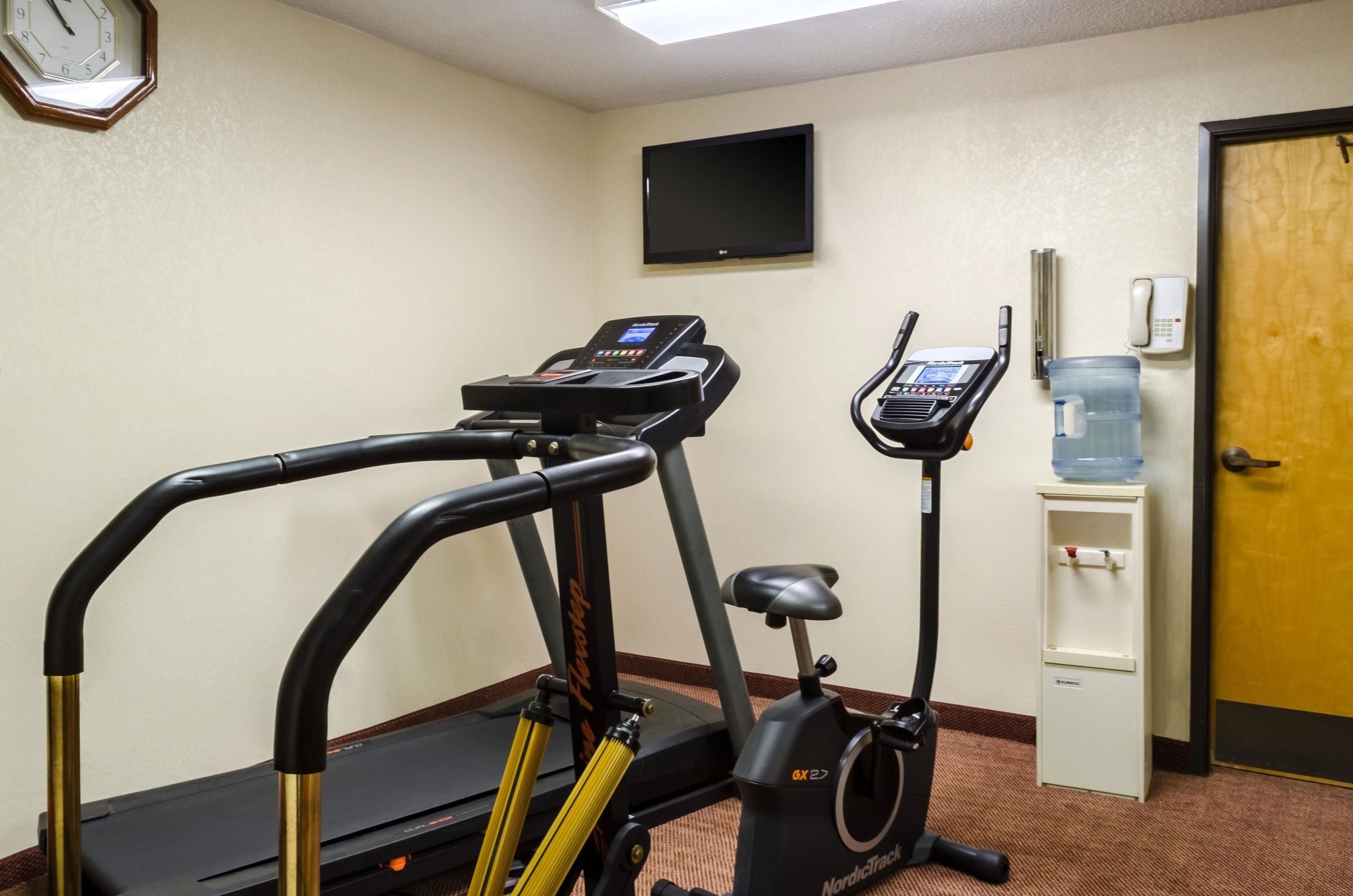 Ein Fitnessraum mit Laufbändern, einem Ergometer, einem Wasserspender, einer Uhr und einem Fernseher an der Wand.
