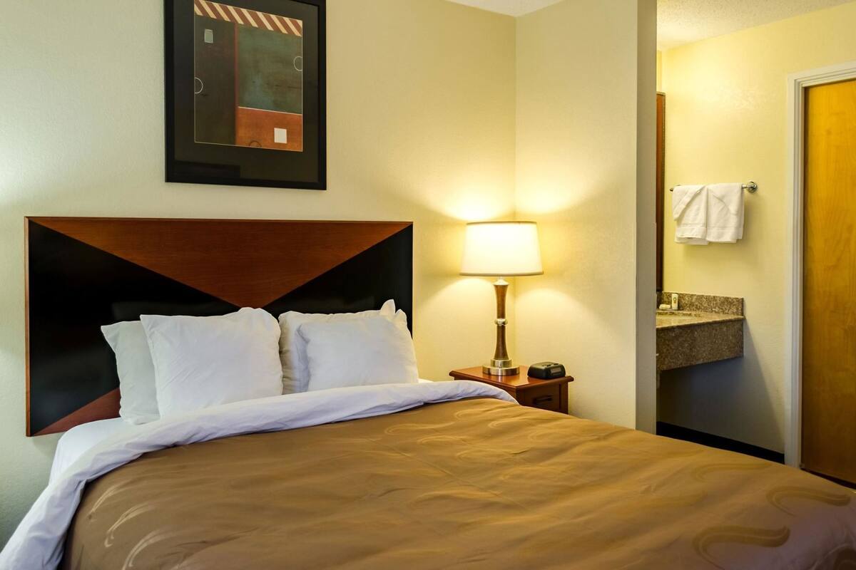 Habitación de hotel con cama, mesita de noche, lámpara, un cuadro enmarcado y espejo de baño.