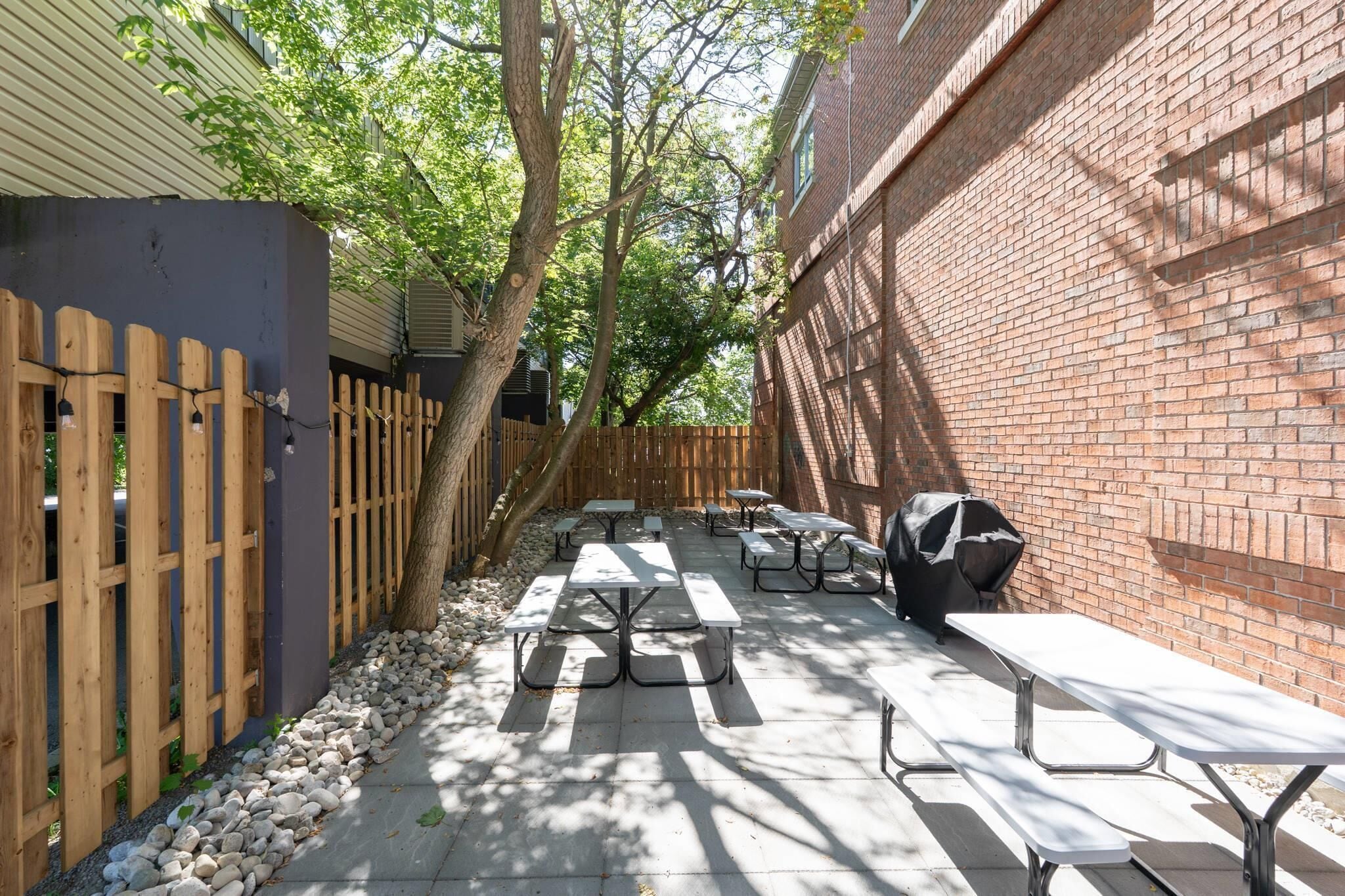 An outdoor patio area with picnic tables and a barbecue grill, surrounded by a wooden fence and a brick wall.
