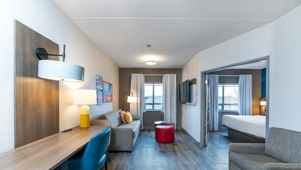 Ein modernes Hotelzimmer mit einem Holztisch, einem blauen Sessel, einem grauen Sofa, einem roten Hocker und einem Bett.
