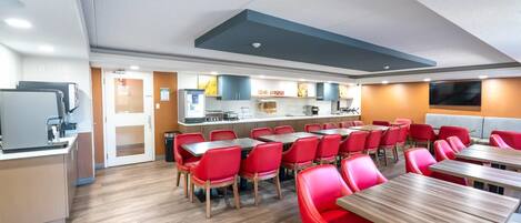 A modern dining area with red chairs and wooden tables, a kitchen in the background, and a flat-screen TV mounted on the wall.