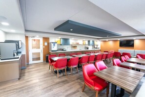 Un espace repas moderne avec des chaises rouges et des tables en bois, une cuisine en arrière-plan et un téléviseur à écran plat fixé au mur.