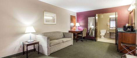 A hotel room with a sofa, a small table with a lamp, a desk with a chair, a bathroom with a toilet and sink, and a mirror.
