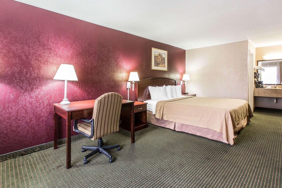 A hotel room with a bed, desk, chair, lamps, and a mirror.