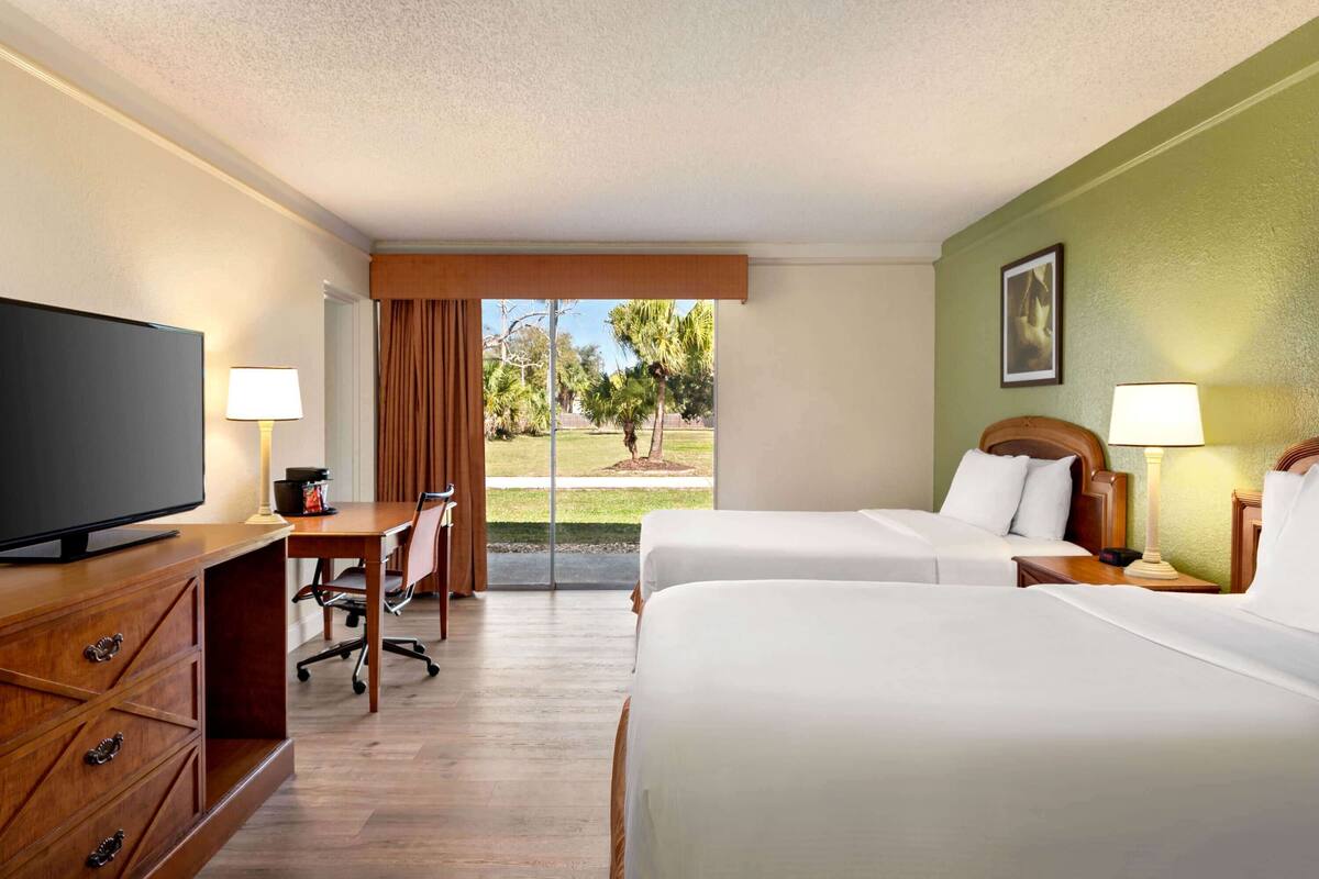 A hotel room with two beds, a desk, a television, and a view of a garden through a glass door.