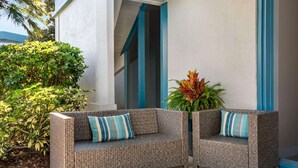 A wicker sofa with striped cushions, positioned on a patio with a potted plant and a building in the background.