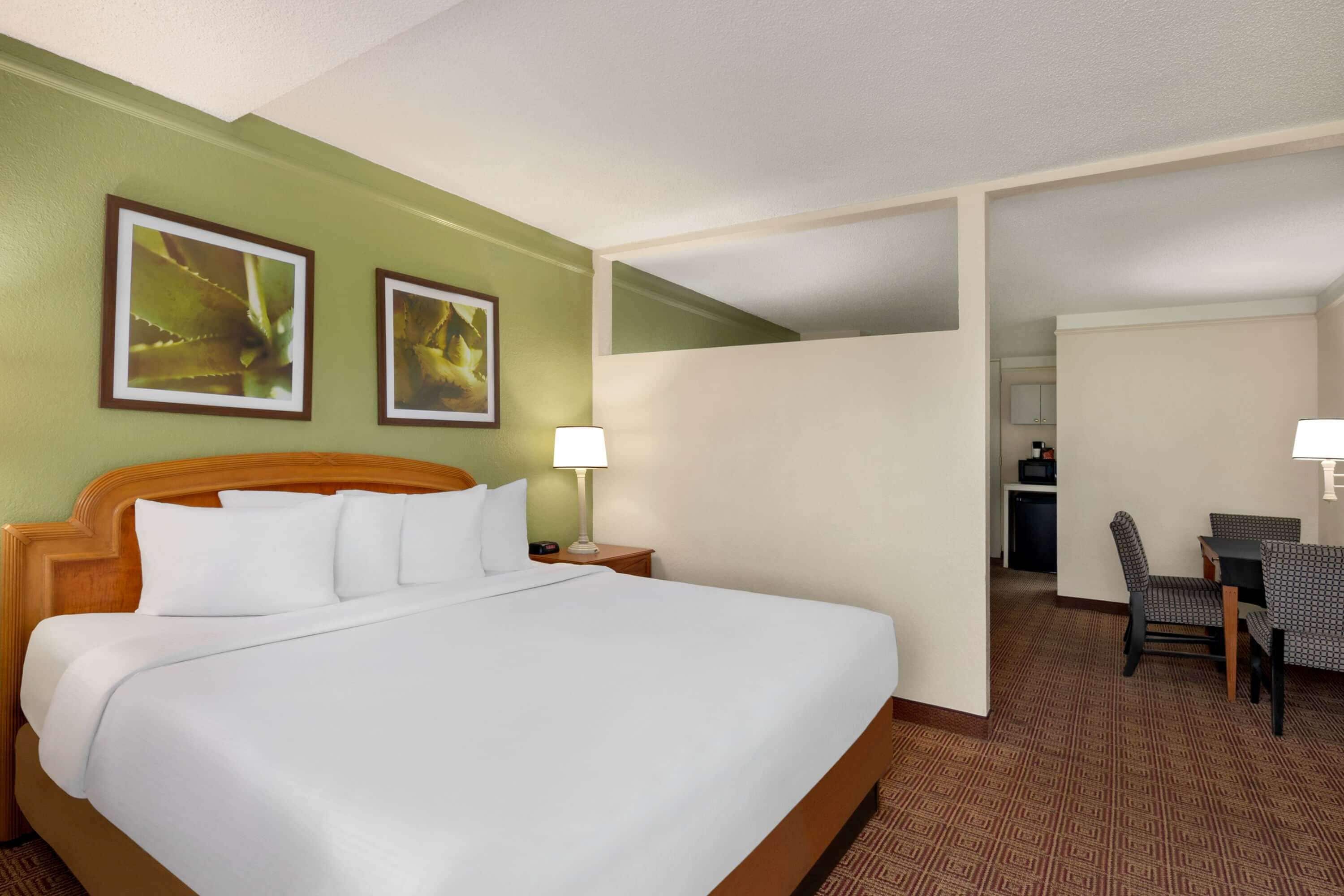 A hotel room with a large bed, two wall-mounted picture frames, a desk with a chair, and a small kitchenette in the background.