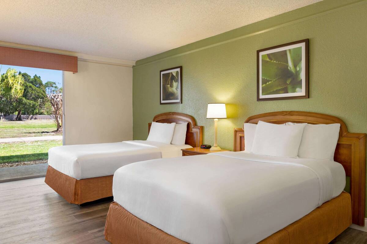 A hotel room with two beds, a window with a view of greenery, and a lamp on the nightstand.