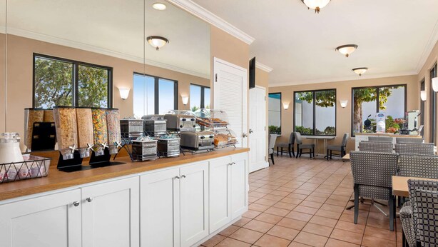 A breakfast bar with various food dispensers and a coffee machine, surrounded by seating areas and large windows.