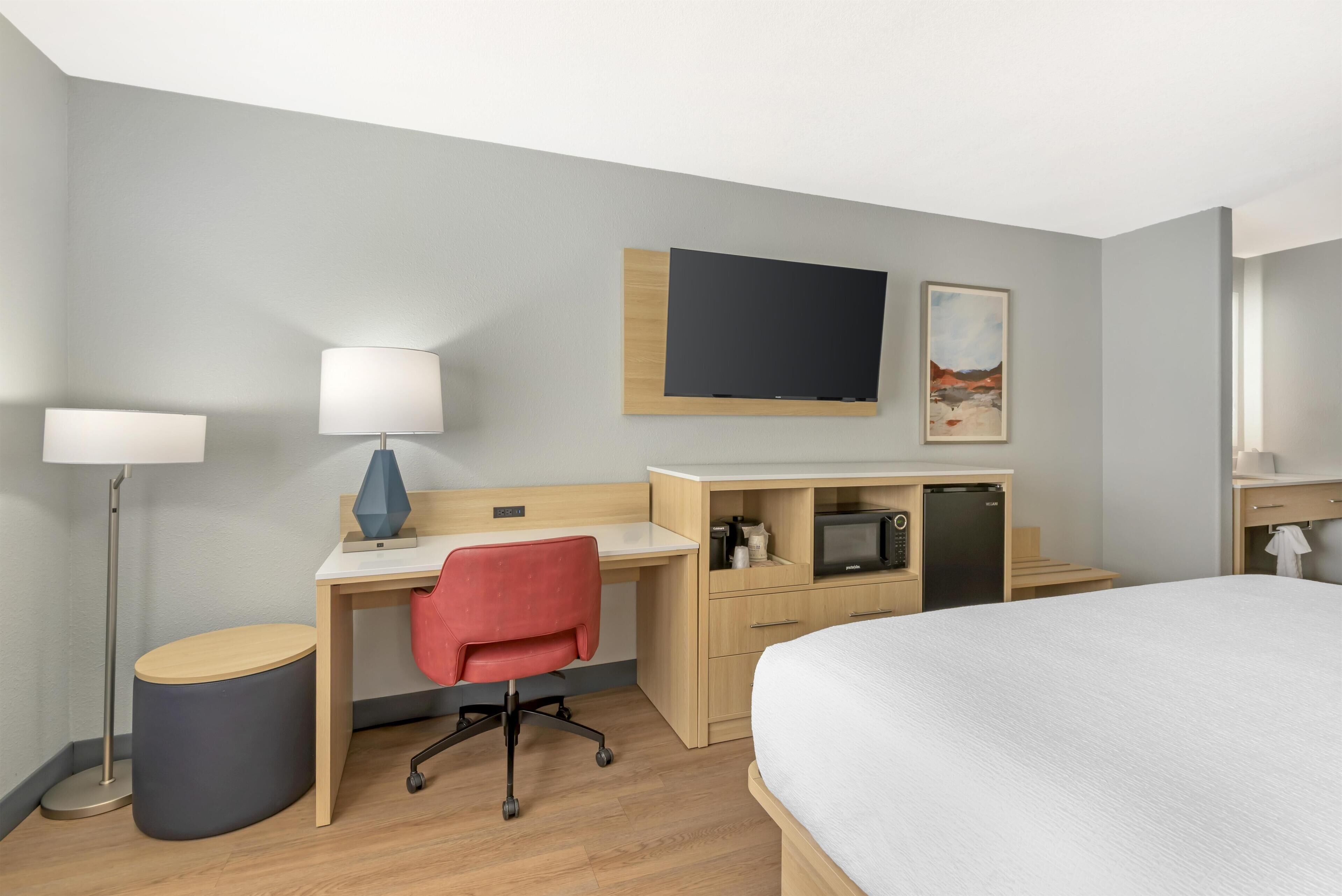 Habitación de hotel con cama, escritorio, silla, lámpara, televisor y una pequeña kitchenette.
