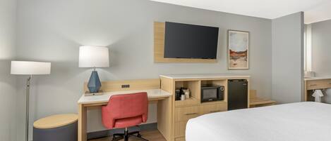 Habitación de hotel con cama, escritorio, silla, lámpara, televisor y una pequeña kitchenette.