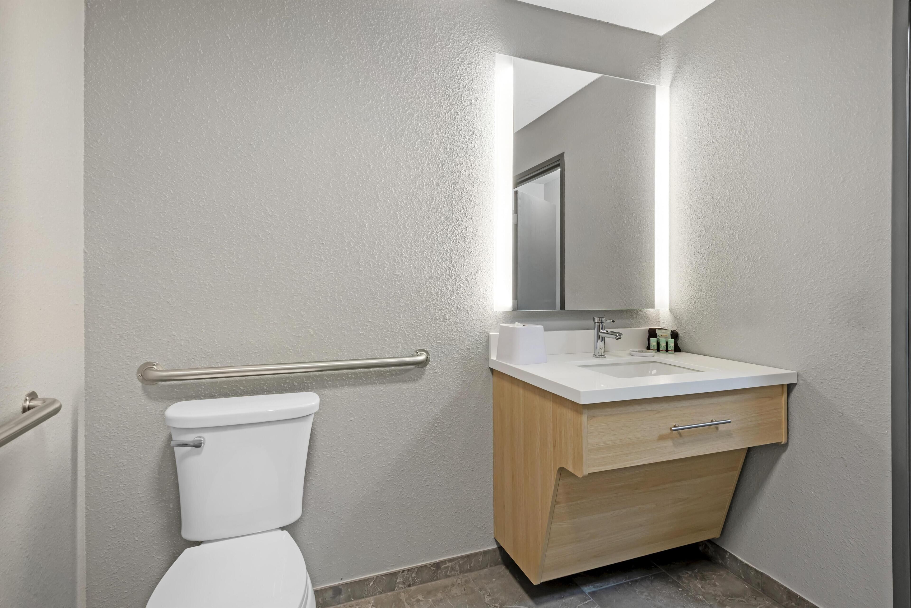 A modern bathroom with a white toilet, a wooden vanity, a mirror, and a handrail.