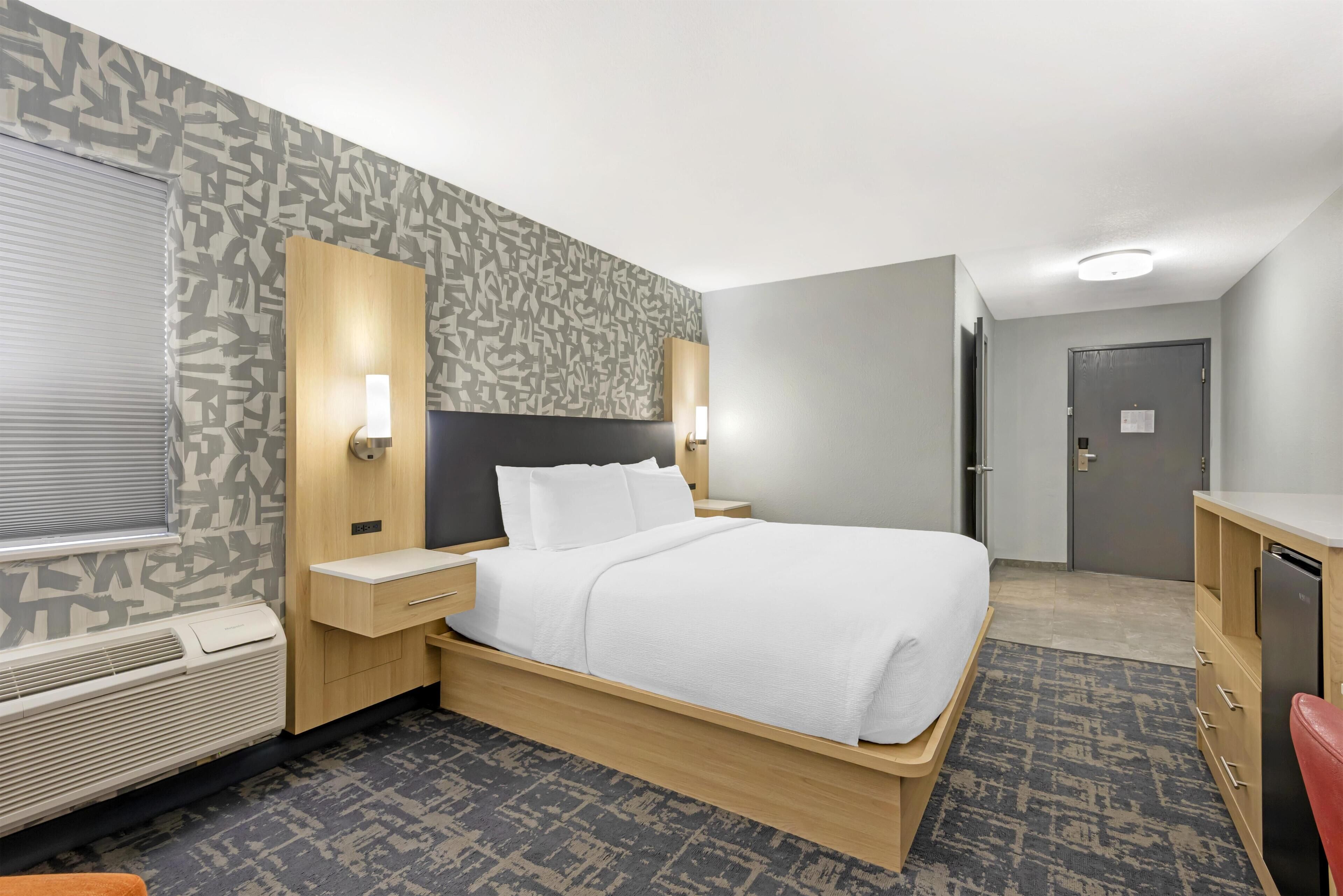 A hotel room with a bed, bedside tables, a desk, a chair, a wardrobe, a heater, and a patterned wall.