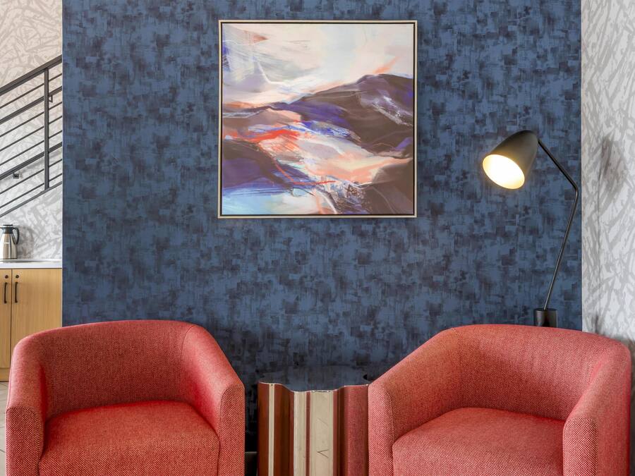 Quarto com parede azul texturizada, um quadro abstrato emoldurado, duas cadeiras vermelhas, um candeeiro de chão preto e um armário de madeira com uma chaleira.