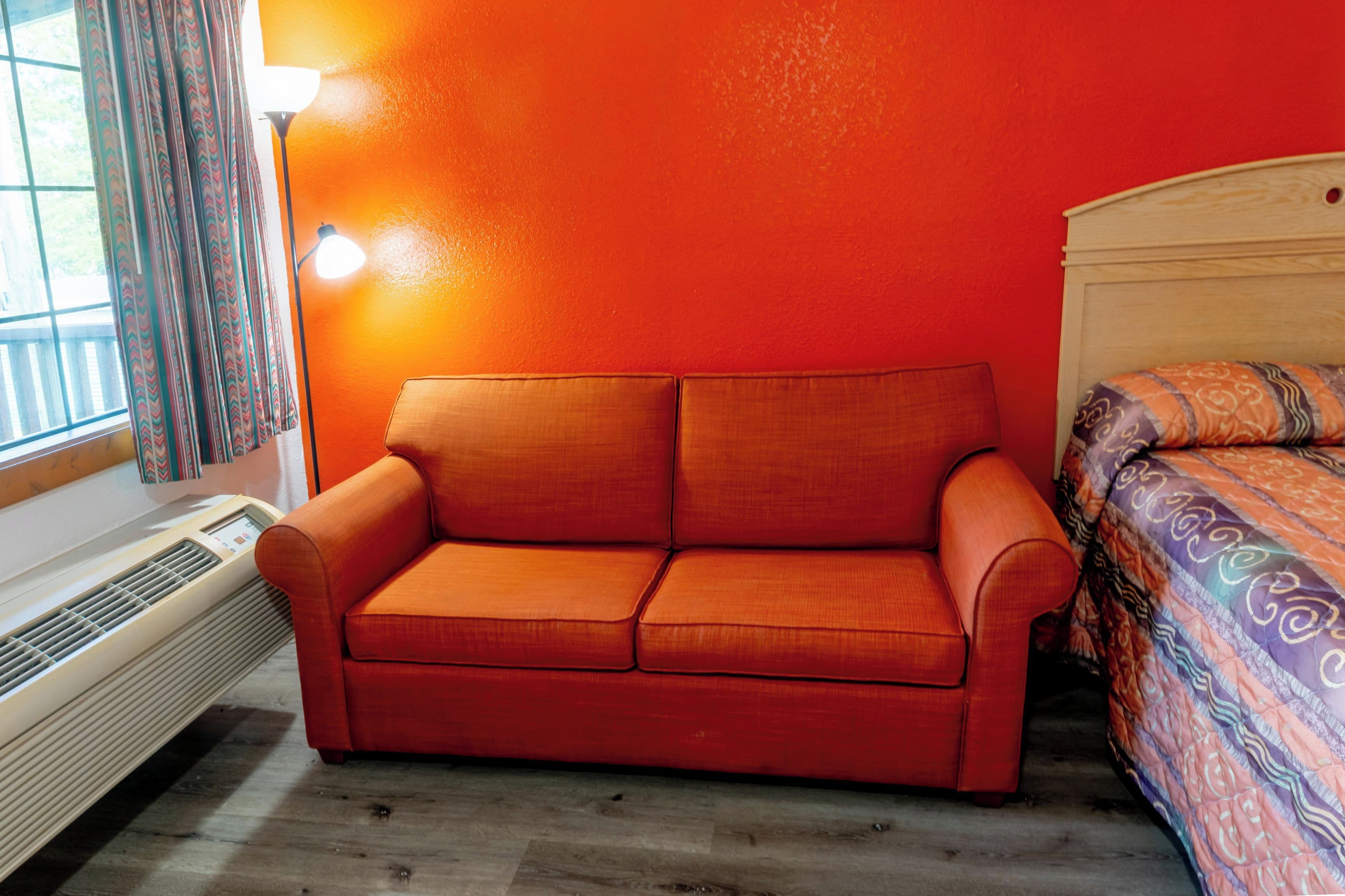 A room with a red sofa, a wooden headboard, a patterned bedspread, and a window with curtains.