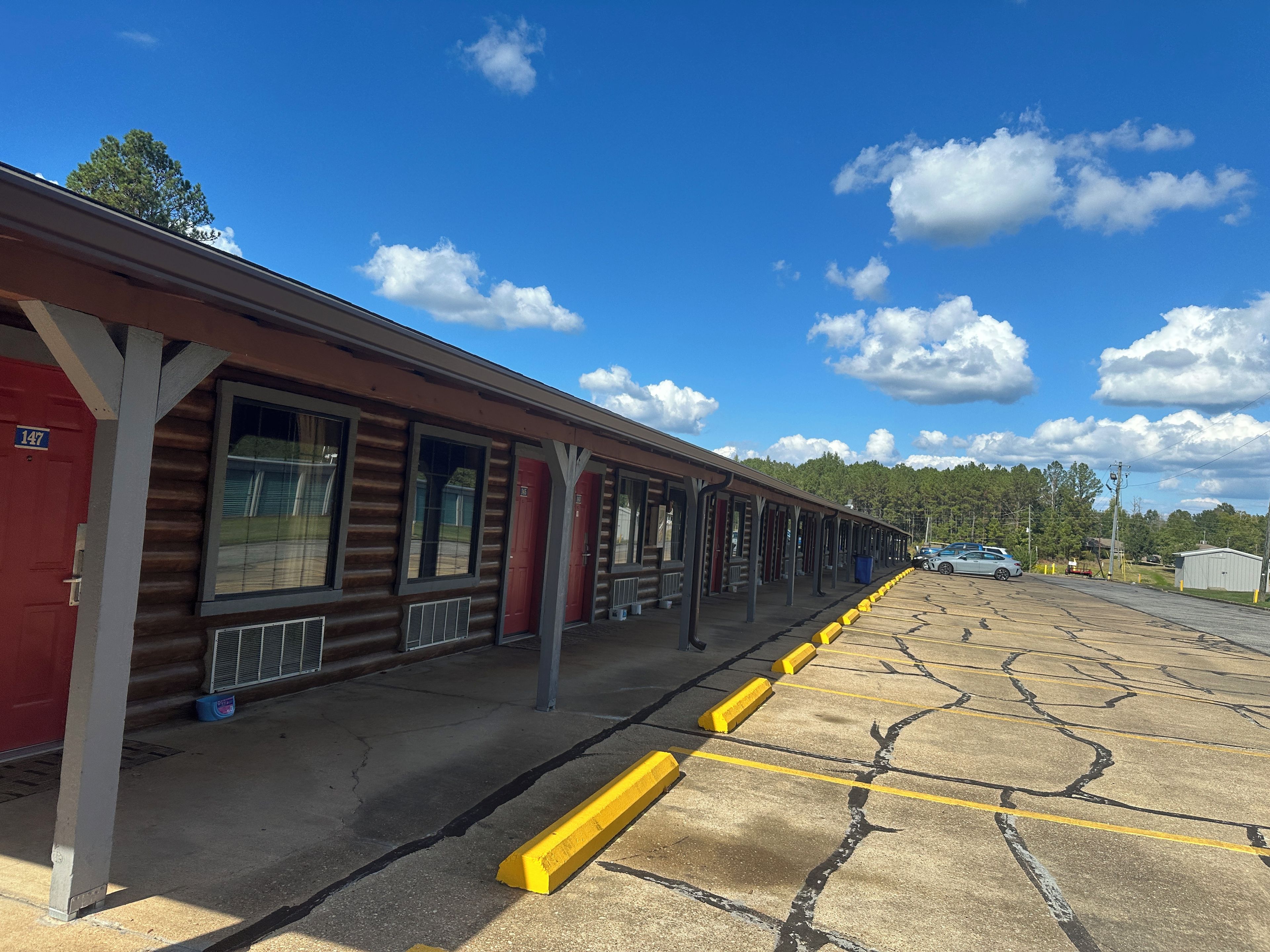 A row of log cabin-style rooms with large windows, a covered outdoor area, and a parking lot with yellow curbs.