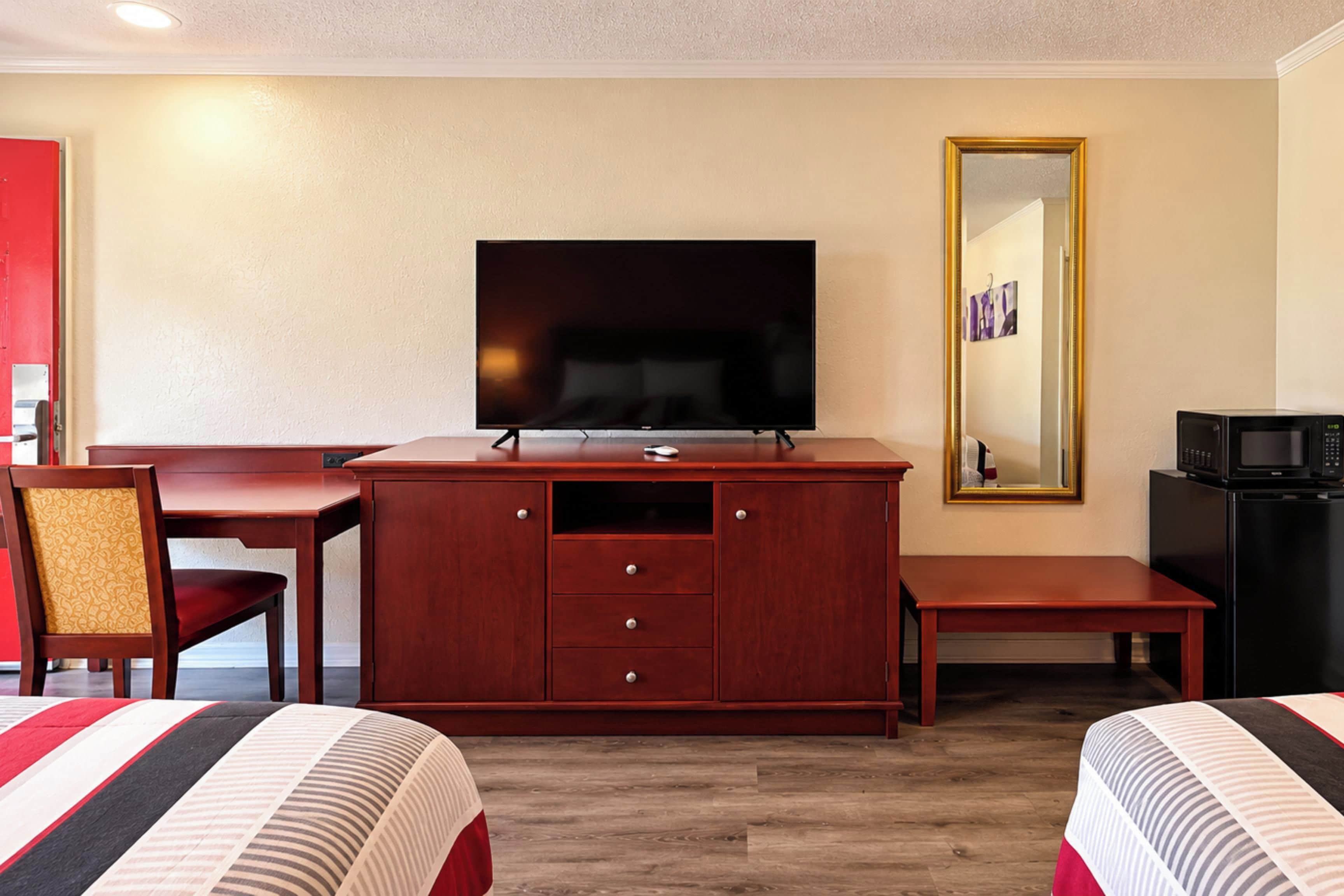 A hotel room with a TV, a desk, a chair, a bed, a microwave, and a mirror.