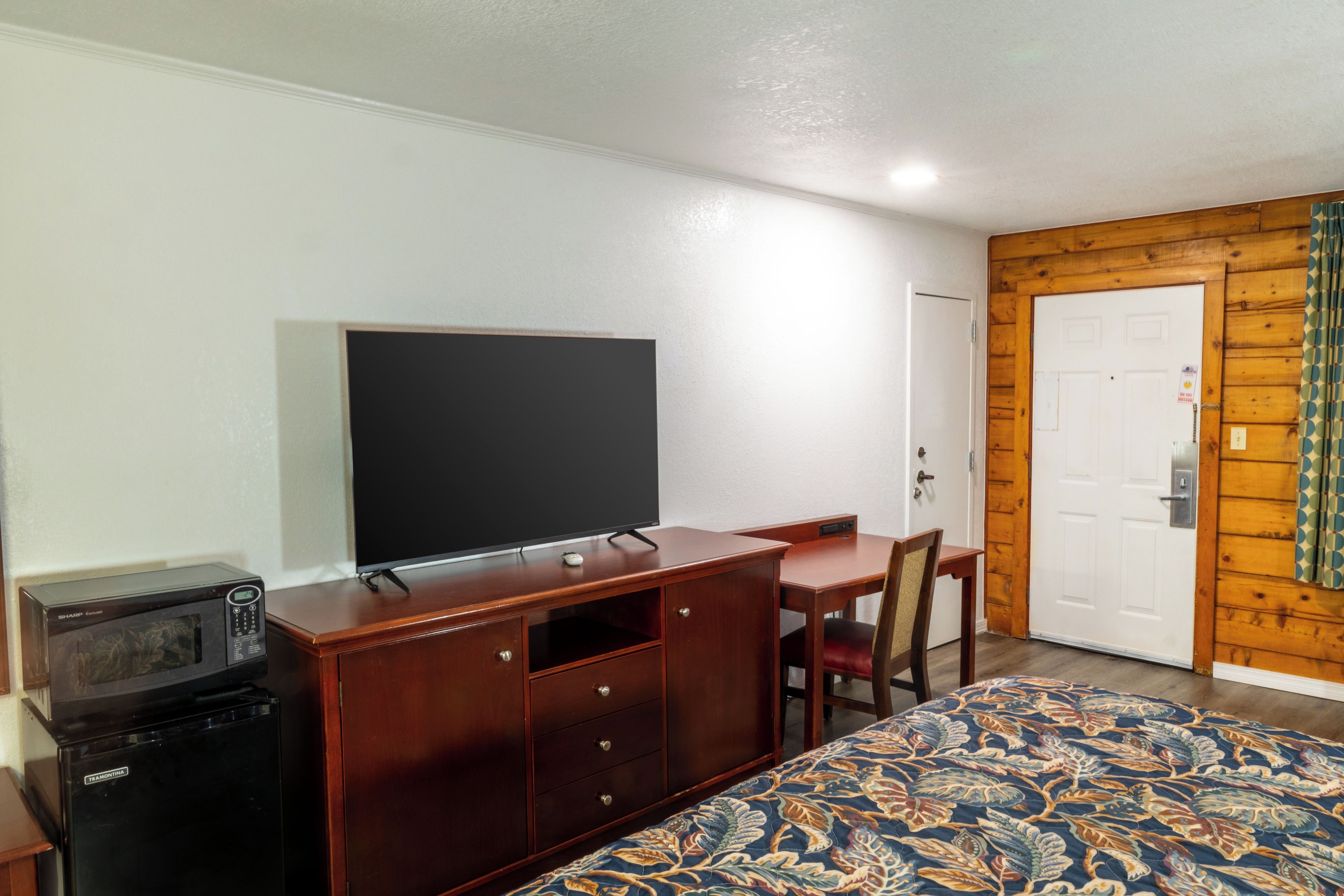 A hotel room with a bed, a television on a wooden dresser, a desk, and a door.