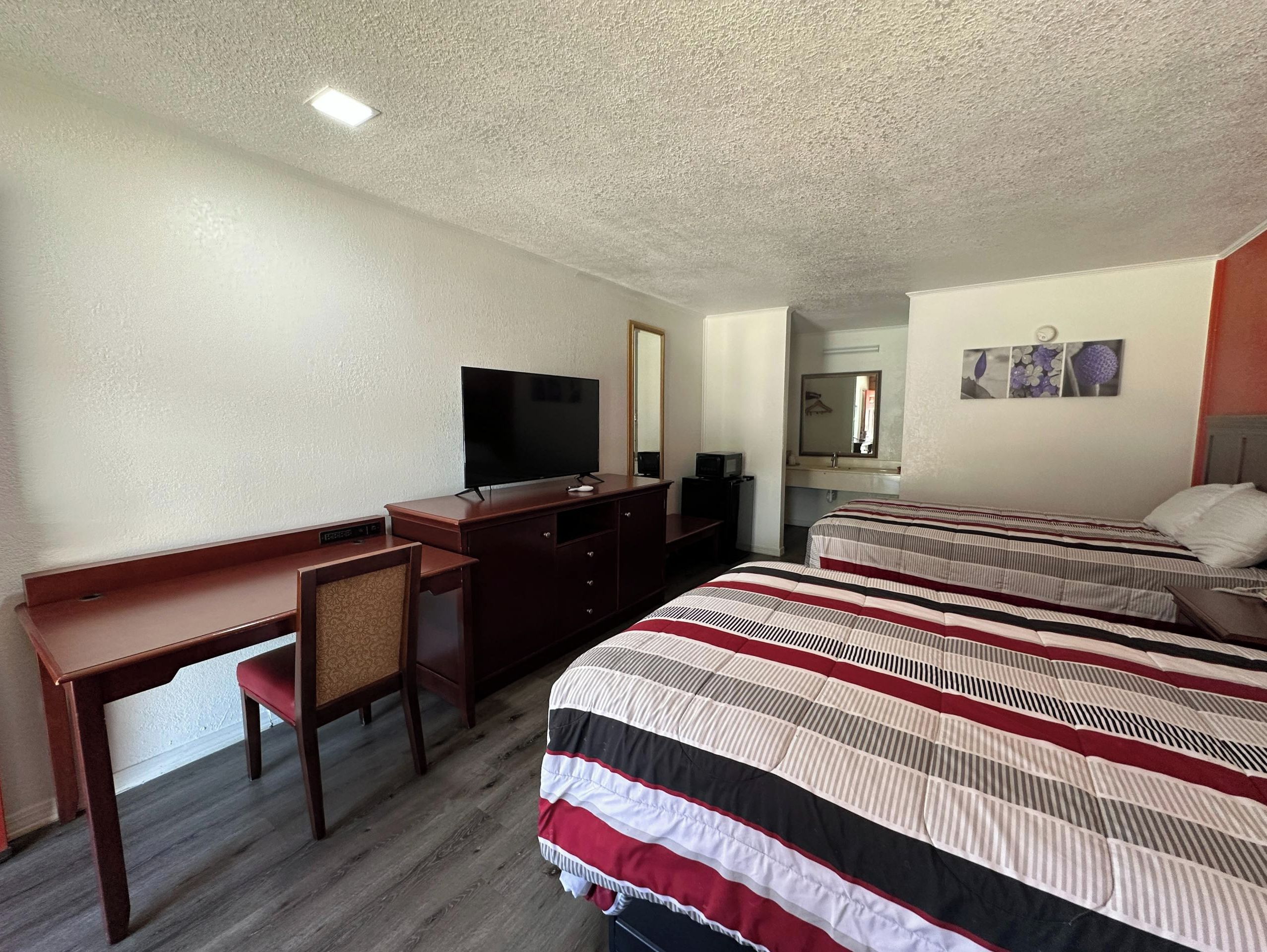 A hotel room with a desk, a television, a bed with striped bedding, a mirror, and a small artwork on the wall.